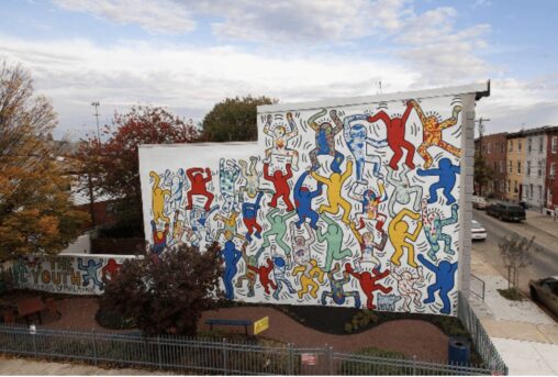 Keith Haring's vibrant outdoor mural depicts several simple human figures in shades of red, yellow, blue, and green that are highlighted in black, overlap, and appear to be moving across a big white wall.