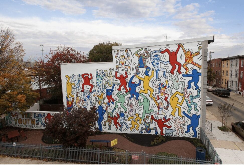 Keith Haring's vibrant outdoor mural depicts several simple human figures in shades of red, yellow, blue, and green that are highlighted in black, overlap, and appear to be moving across a big white wall.