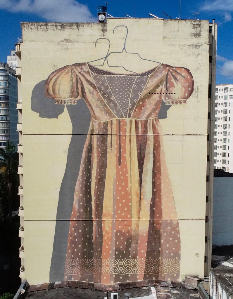 What Remains by Hyuro, Large-scale mural on the side of a building depicting an empty, old-fashioned dress hanging on a wire hanger, painted in muted earth tones against a pale wall, with no body inside, evoking absence, memory, and what is left behind.